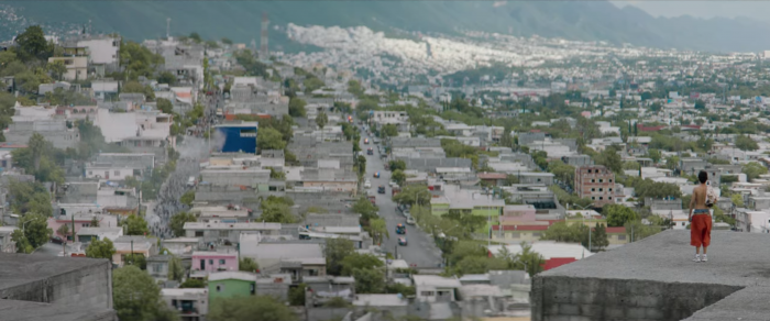 Ulises, sin player, encima de un edificio de concreto, mirando el resto de la ciudad con casas de concreto. Una calle se observa llena de personas